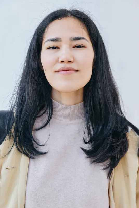 Young woman with long dark hair in a beige jacket and sweater, neutral expression against a light background, casual calm look highlighting skin tone and mood