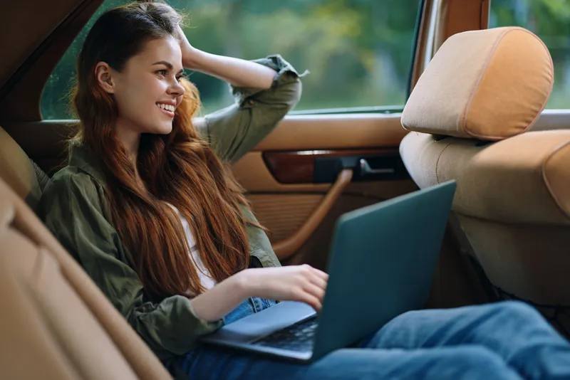Young woman working on a laptop in a car, smiling and relaxed, showcasing a casual outfit and warm autumn colors.