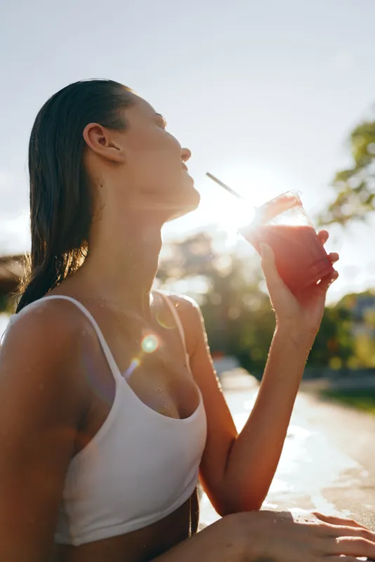 Young woman in a white tank top sits outdoors on a sunny day, drinking juice from a cup, enjoying warm light and a relaxed lifestyle moment, candid and fresh, natural backdrop, gentle breeze,