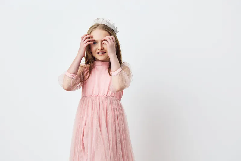 A joyful young girl wearing a pink dress with sheer sleeves and a floral crown, expressing happiness in front of a minimalistic background