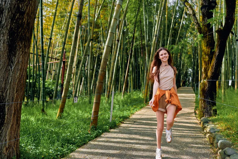 Woman walking on a bamboo path in a green forest, smiling and wearing casual summer outfit, long hair flowing with sunlight filtering through tall bamboo stalks along a gravel trail.