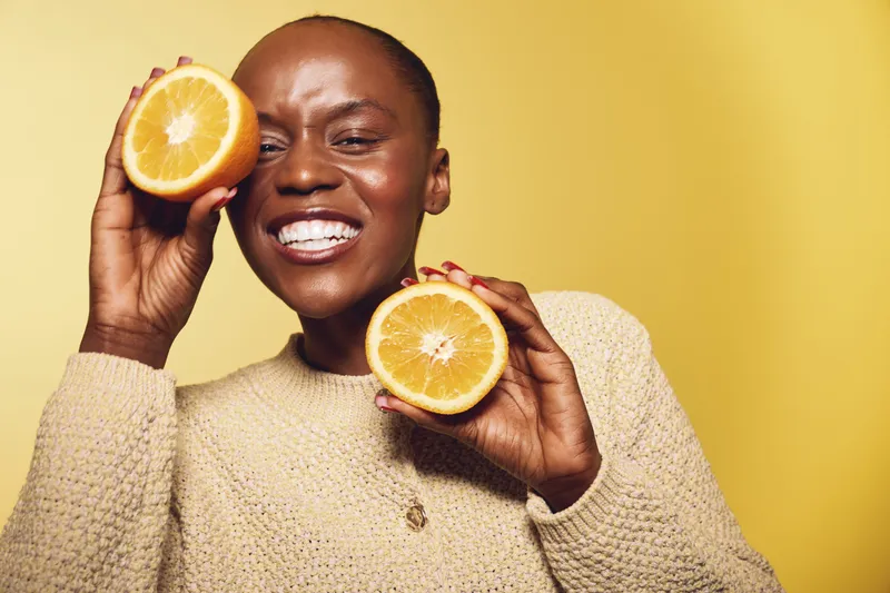 Happy African woman with dark skin holds two halves of fresh orange fruit near face smiling. Bright yellow background, casual sweater, concept of healthy lifestyle and natural vitamins.