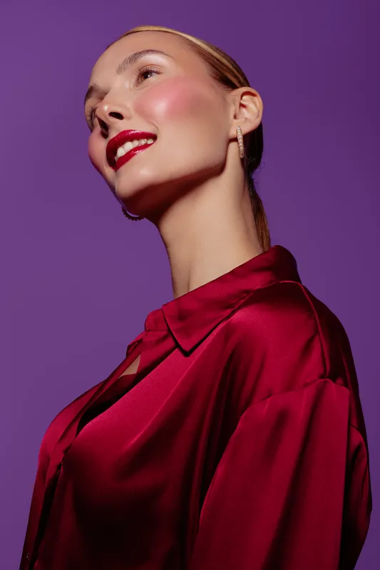 Young woman with glossy red lips and shiny skin wears red satin blouse against purple background. Portrait with elegant makeup and smooth hairstyle. Concept of beauty, fashion, and glamour.