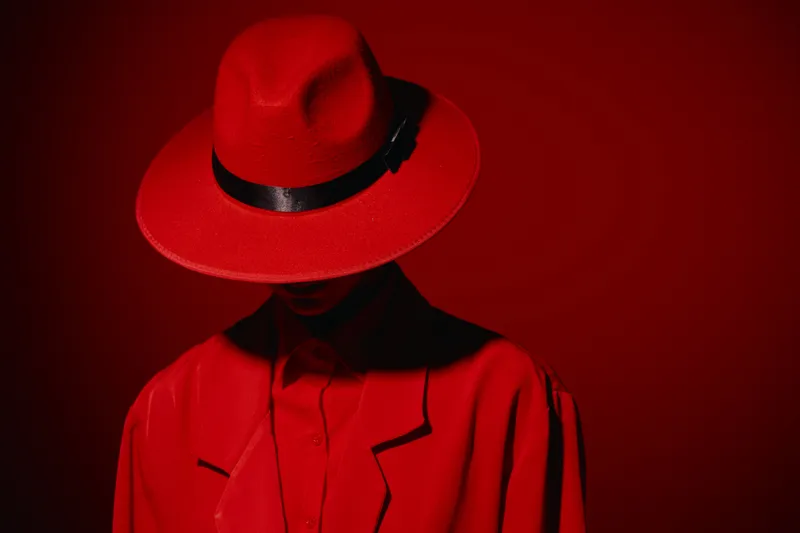 Person dressed in a red hat and matching coat posing against a vivid red background. Dramatic lighting creates a mysterious vibe for fashion or beauty concept.
