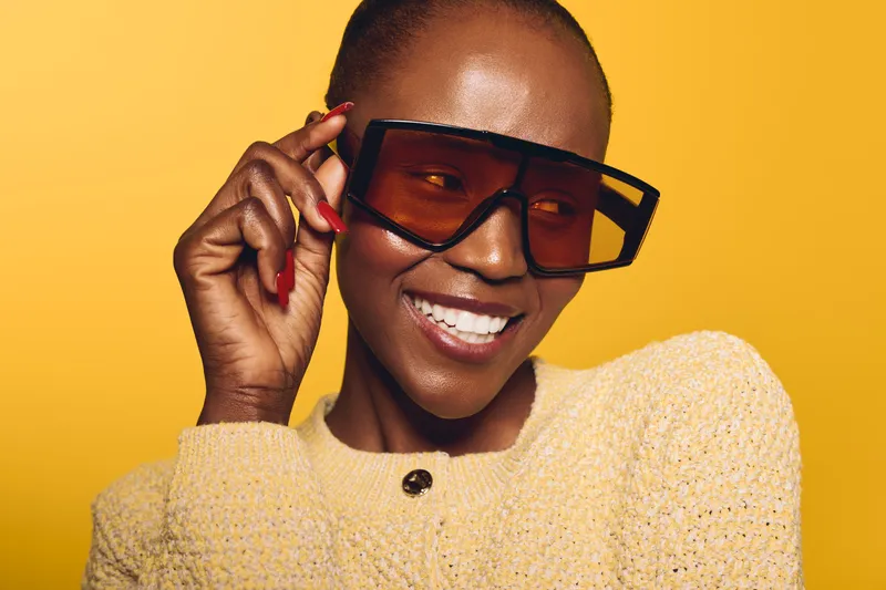 Confident dark skinned African woman with short hair wearing oversized sunglasses and a light sweater smiles against yellow background. Concept of fashion, beauty, and style with joyful mood.