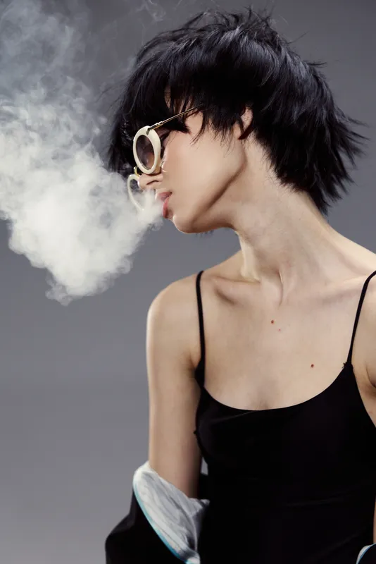 Profile of a woman with short black hair exhaling smoke wearing sunglasses and a black top