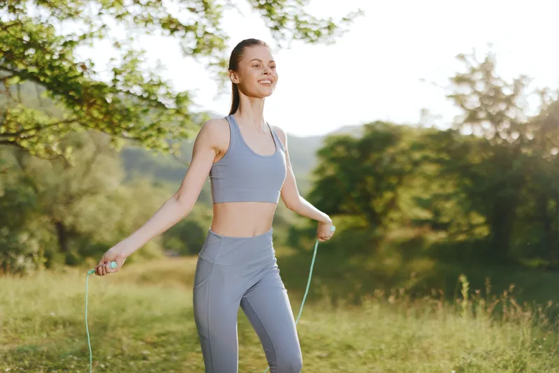 Young woman exercising outdoors in gray sportswear, smiling while skipping rope in a sunny green field, promoting a healthy lifestyle and fitness