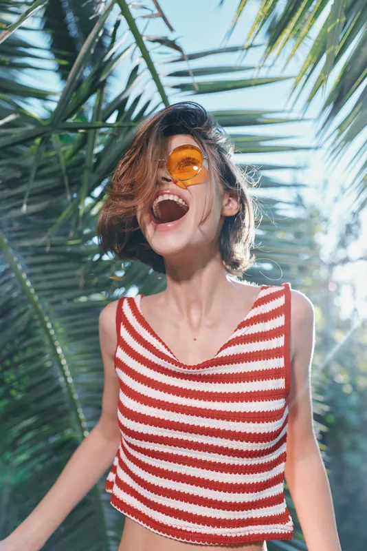 A joyful young woman wearing a red and white striped crop top and orange sunglasses laughs outdoors among tropical plants, radiating carefree summer energy.