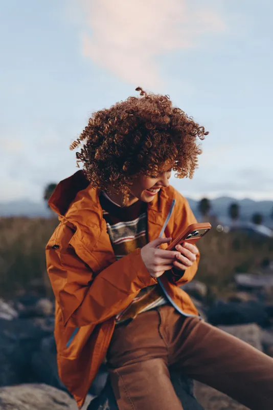 Woman lifestyle outdoors, curly hair smiling, orange jacket, using smartphone on a hill at sunset, warm light, soft breeze, distant mountains blurred, relaxed moment, in countryside