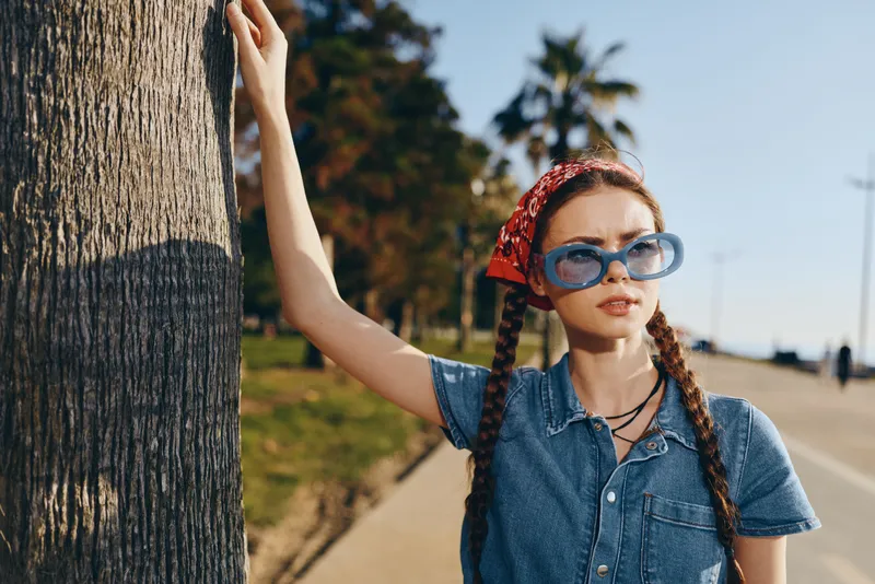 Lifestyle woman in modern boho-western denim street style wearing blue sunglasses and red bandana with braids, outdoor sunny urban background, film color effect.