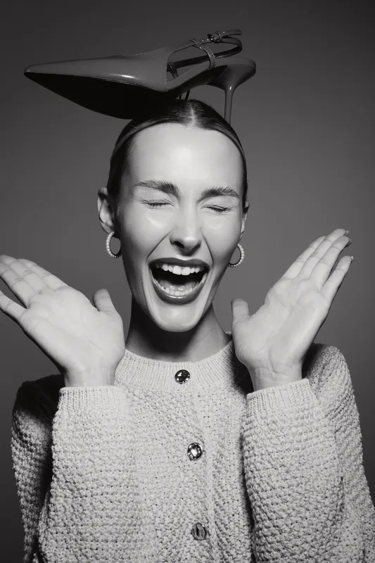 Young white woman with pale skin and closed eyes laughs holding hands near face wearing stylish sweater and shoes on head.