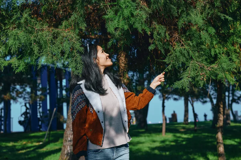Woman in a brown suede jacket and cozy sweater stands in a sunny park, reaching toward leafy branches, enjoying nature and outdoor light in a peaceful moment
