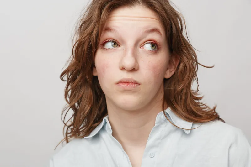 Young woman with curly hair looks sideways with thoughtful expression wearing light blue shirt. Studio portrait on white background. Concept of doubt, hesitation, and contemplation.