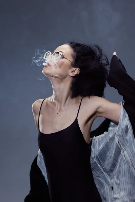 Elegant woman with glasses exhaling smoke, wearing black dress and light jacket, exhibiting a relaxed and artistic vibe.