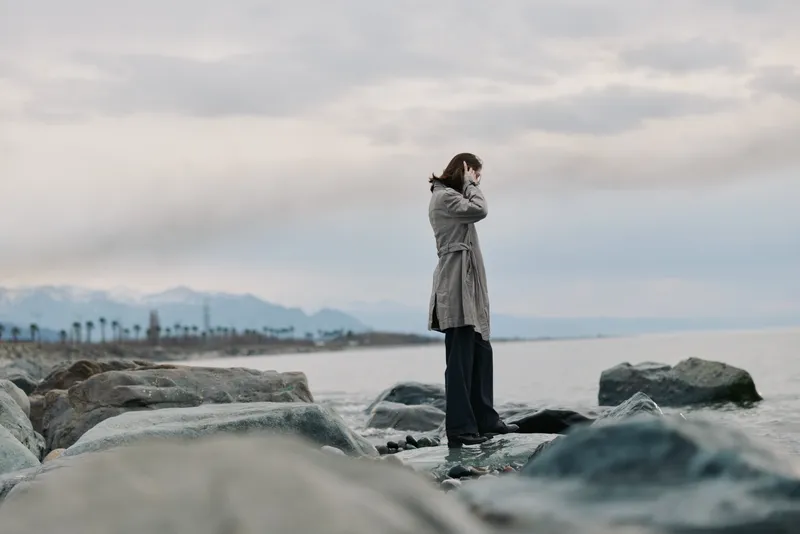 A thoughtful woman in a coat stands on rocky shore facing calm sea under overcast sky. The distant mountains and palm trees add to the serene, contemplative coastal atmosphere.