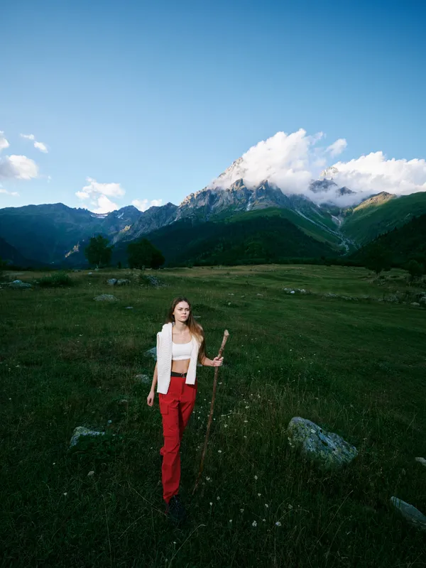 Mountain landscape, outdoor scene with a woman wearing red pants standing in a meadow, distant peaks, clear sky, rugged terrain and calm atmosphere, ideal for travel, exploration and nature