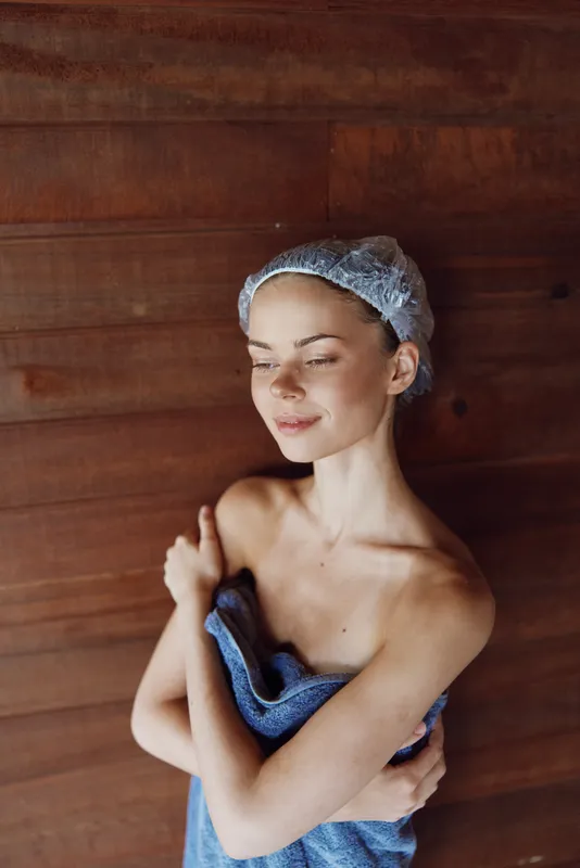 Relaxed woman in shower cap wrapped in blue towel, standing against wooden wall, enjoying serene moment with closed eyes, soft natural lighting, calm and peaceful mood.