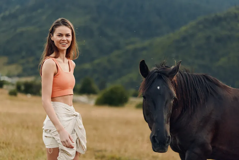 A cheerful woman stands in a sunny field beside a horse, wearing a coral top and light shorts. She smiles warmly, enjoying a calm outdoor moment.