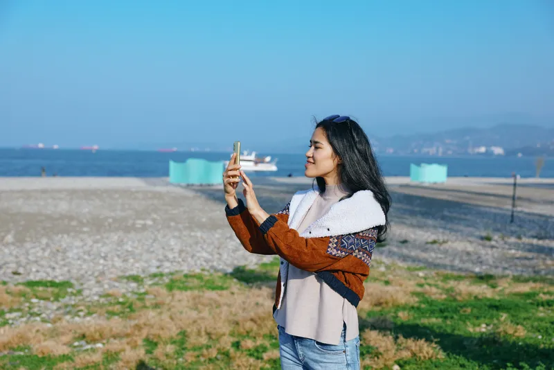 Selfie moment on the shoreline, a relaxed woman frames a sunny beach scene with a smartphone, casual coat and breeze in her hair, capturing coastal lifestyle and travel vibes by calm water.