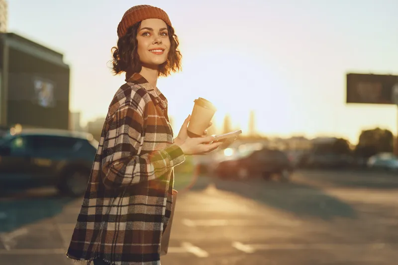 Woman with coffee and smile in urban street at golden hour, wearing beanie and plaid coat. Candid lifestyle portrait capturing authenticity, mindful living and emotional storytelling.