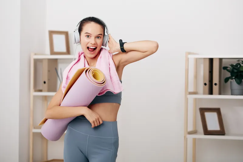 Woman fitness enthusiast in a bright home studio with headphones, holding a rolled yoga mat and wearing a sports bra and leggings; energetic smile, minimal shelves and plants in the background.