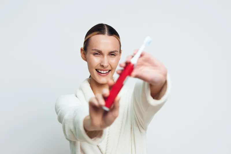 Smiling woman with a toothbrush extended toward the camera, showcasing a dental care routine. Bright, neutral background. Concept of oral hygiene, wellness, and self care. Warm lighting.