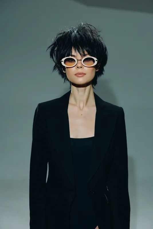 Stylish woman with short black hair wearing white sunglasses and a black blazer posing confidently against neutral background