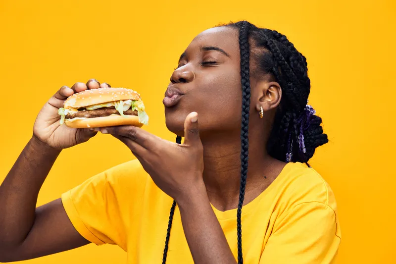 Happy young woman with braids enjoying a delicious burger against a vibrant yellow background, showcasing her love for fast food in a lively and playful manner