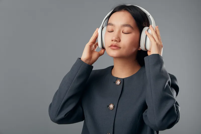 Young woman with closed eyes wearing headphones and dark jacket listening to music or audio with focus against grey background. Relaxed girl enjoying sound and calm moment indoors.