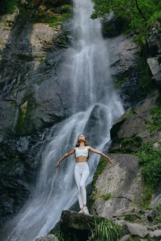 woman enjoying waterfall outdoors, standing on rock with arms outstretched, dressed in white fitness clothes, surrounded by lush green nature and flowing water.