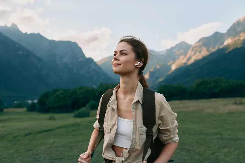 A young woman with earbuds and a backpack enjoys a mountain landscape during a hike. She looks content and relaxed in casual outdoor clothing under a cloudy sky.