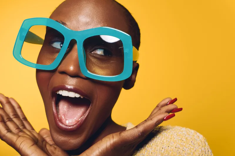 Excited African woman with dark skin wearing oversized blue glasses and yellow sweater poses against yellow background. Concept of joy, fashion, beauty, and expressive emotions.