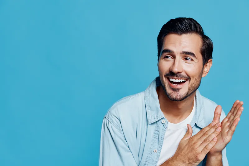 Happy smiling young man with short hair wearing a light blue shirt, sitting against a solid blue background, looking to the side, expressing enthusiasm and joy.
