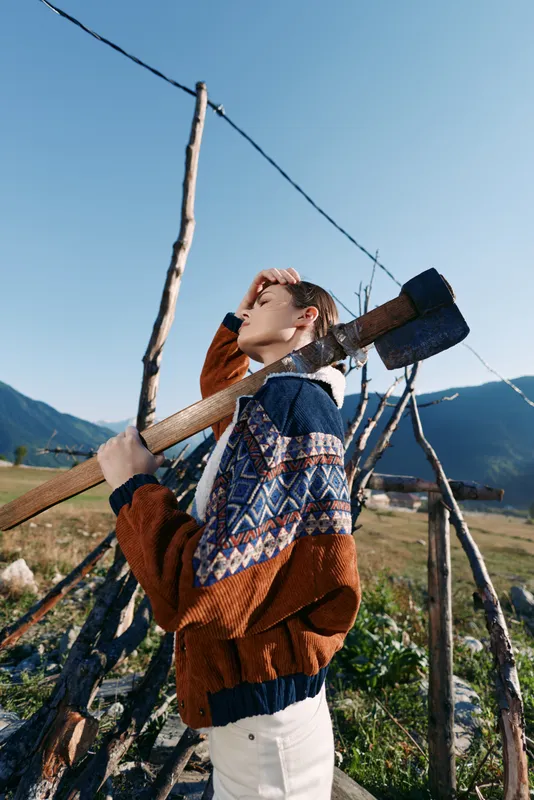 woman with axe on shoulder in countryside mountains wearing patterned sweater outdoors by rustic fence, portrait of young female in sunny autumn field for rural lifestyle and manual work