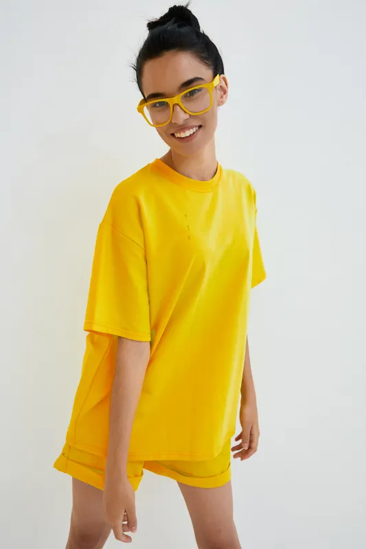 smiling woman wearing yellow oversized t-shirt and matching shorts with trendy yellow glasses against white background, happy expression and casual summer style