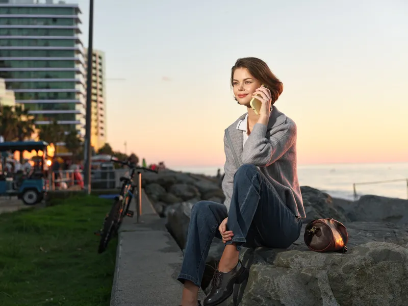 A thoughtful woman sits on a rocky waterfront at sunset, talking on a smartphone. She wears smart casual attire and a relaxed pose, conveying calm connection and confidence.