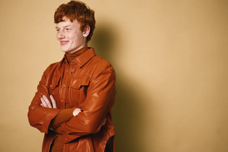 Young man with red hair wearing brown leather jacket and turtleneck posing with arms crossed on beige background studio. Smiling confident male portrait with casual look.