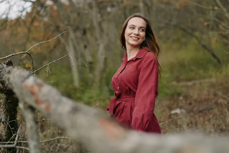 A candid, authentic portrait of a woman in a red coat standing in a rural park, with soft natural light, sincere smile, and subtle texture in the clothing.