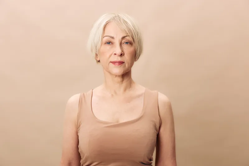 Portrait of an elegant mature woman with short blonde hair wearing a beige tank top, standing against a neutral background. Concept of confidence, aging, and natural beauty.