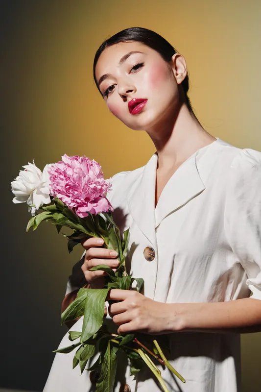 Young woman with bright makeup and bold red lips holds pink and white flowers wearing a modern white outfit. Creative beauty and fashion portrait on warm background.