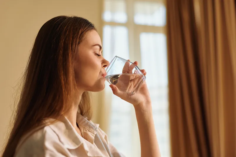 woman drinking water from a glass at home in profile during morning light, hydration and relaxation captured in portrait style, healthy lifestyle and calm domestic routine