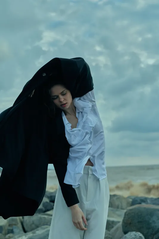 A woman on a windy beach draped in a black cloak over a white shirt and flowing pants, gazing downward amid a moody stormy sky and rocky shoreline at dusk.