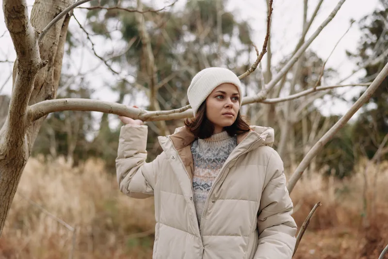 Winter scene with a woman in a beige puffer coat wearing a beanie, standing among leafless trees in a chilly park, creating a calm, candid outdoor portrait.