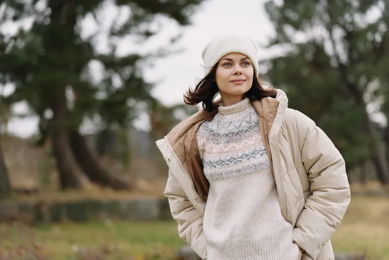 Woman in a warm coat and knitted sweater stands outdoors in a park during chilly weather, wearing a beanie with a calm expression and soft natural light, capturing a cozy seasonal mood