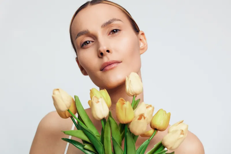 A young woman with clear skin holds a bouquet of yellow tulips close to her face against a plain white background. Concept for natural beauty, spring, and freshness.