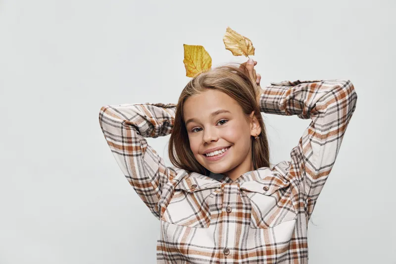 Girl posing and smiling with autumn leaves holding behind head for playful mood with checkered shirt on white background. Child, portrait and casual style for fall season happiness.