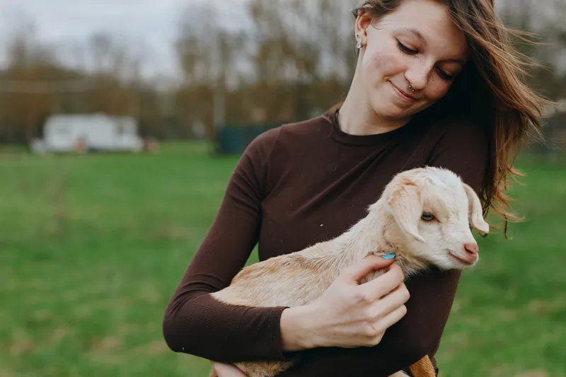 A woman cradles a gentle goat kid in her arms in a sunny country field, capturing a warm lifestyle moment of bonding, care, and rural simplicity.