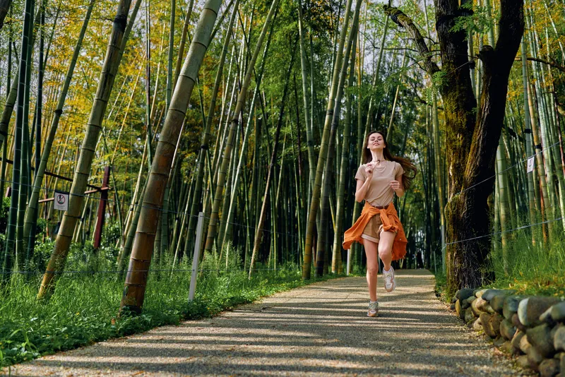 Woman jogging along a gravel path through a sunlit bamboo forest, runner in casual sportswear with sweater tied at waist, enjoying nature and outdoor exercise on a trail.