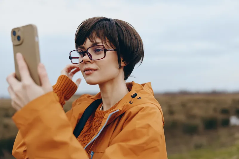Woman lifestyle outdoors, taking selfie with smartphone, orange jacket, glasses, holding phone near face, looking at camera, casual portrait, open landscape background, windy day, natural light