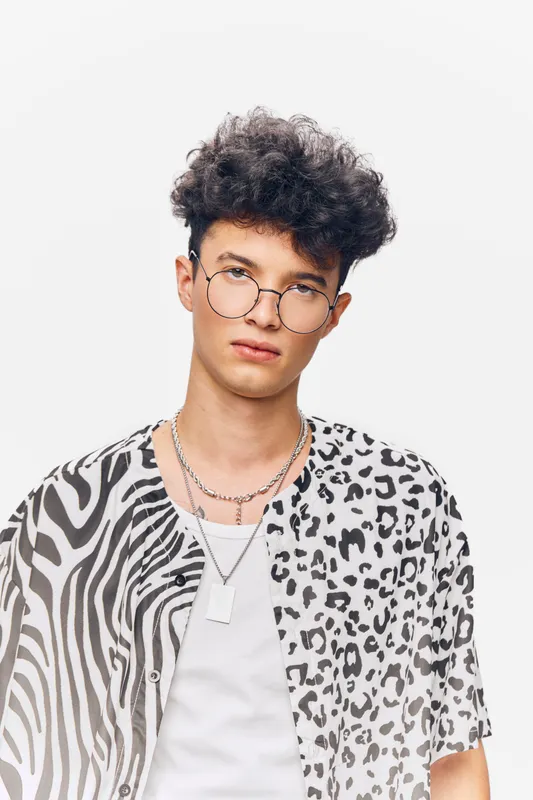 young man with curly hair wearing round glasses and mixed animal print shirt standing against white background in casual style